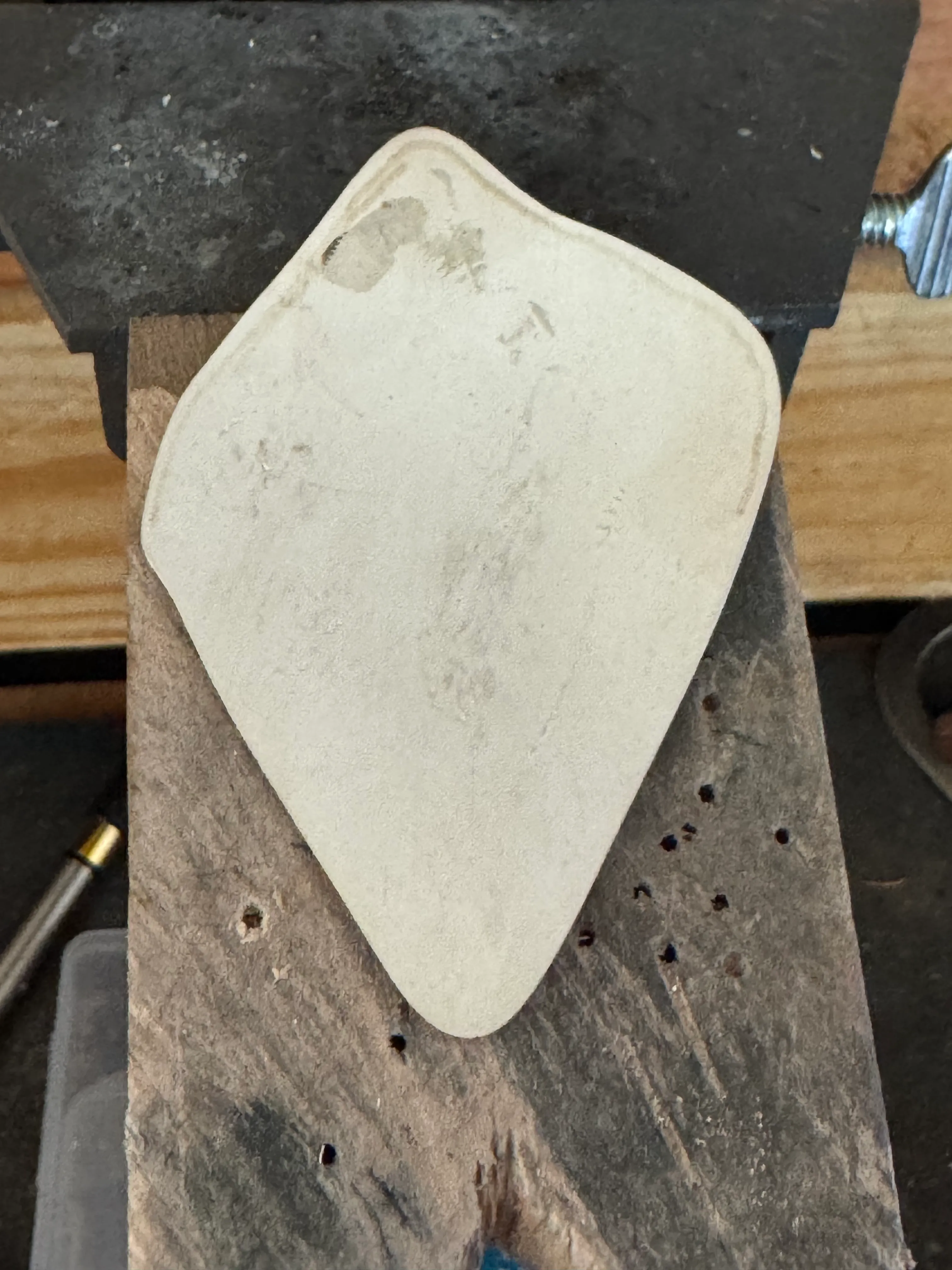A flat, matte-white silver blank roughly sawn to a teardrop shape, resting on a wooden block in a vise, with no markings on it at all.
