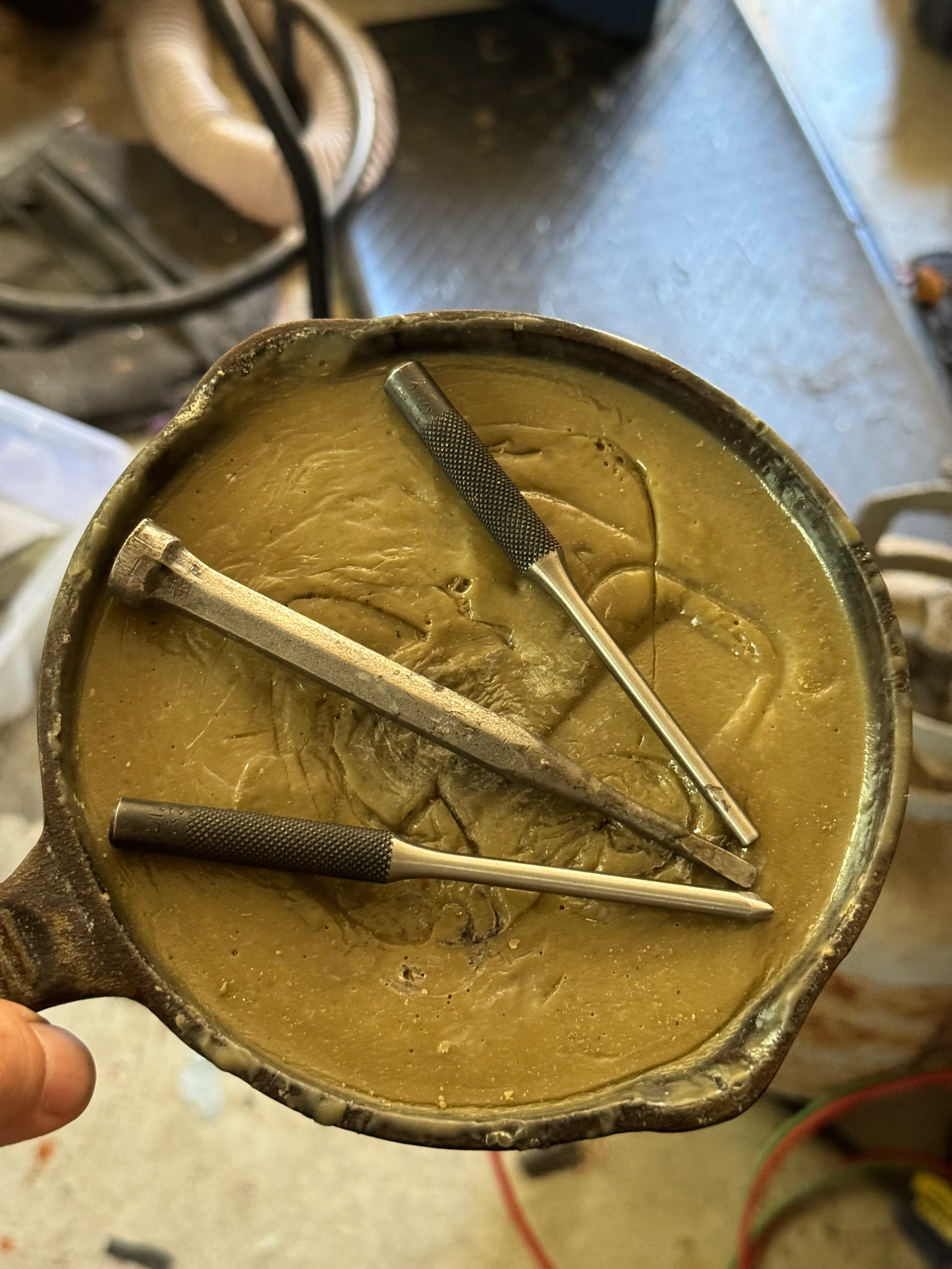 A small cast iron skillet filled with olive-colored chasing pitch, with three hand-made steel punches lying on its surface.