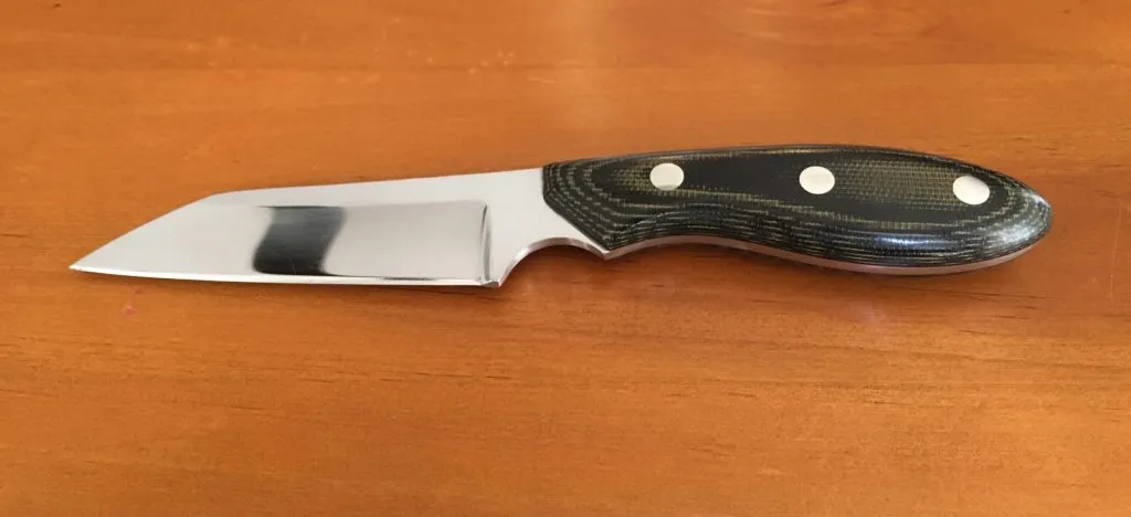 A small finished knife lying on a wooden tabletop: mirror-polished blade with a straight edge and angled tip, and a green-and-black micarta handle set with three pins.